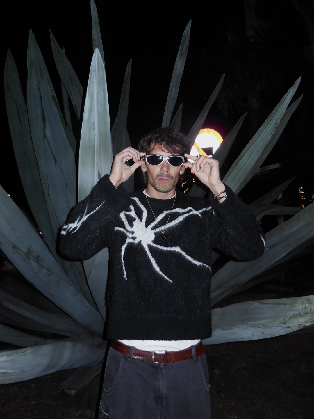 Artist in white sunglasses and spider knit against agave leaves at night